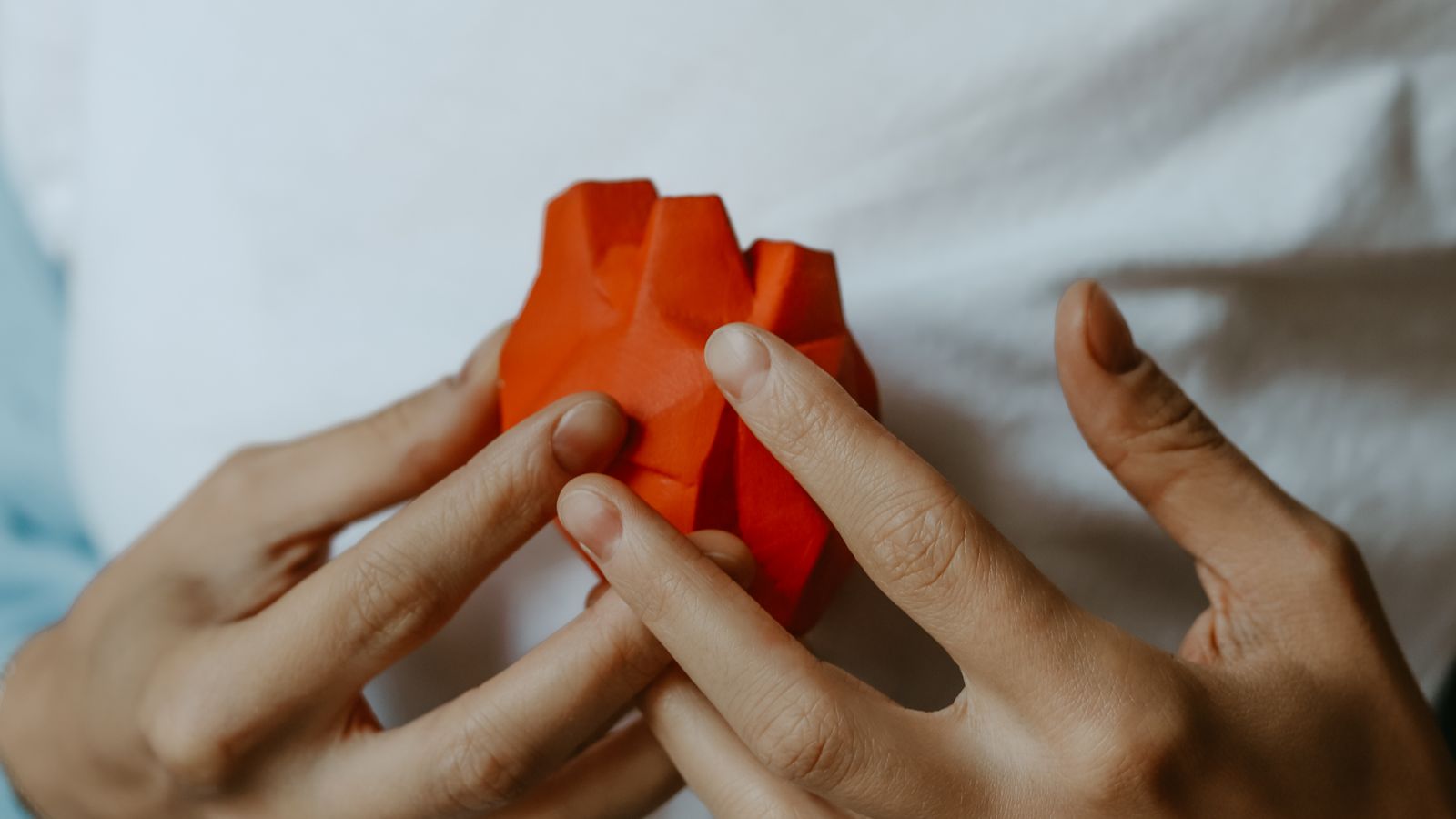Hands holding an orange folded-paper heart model against a white top.