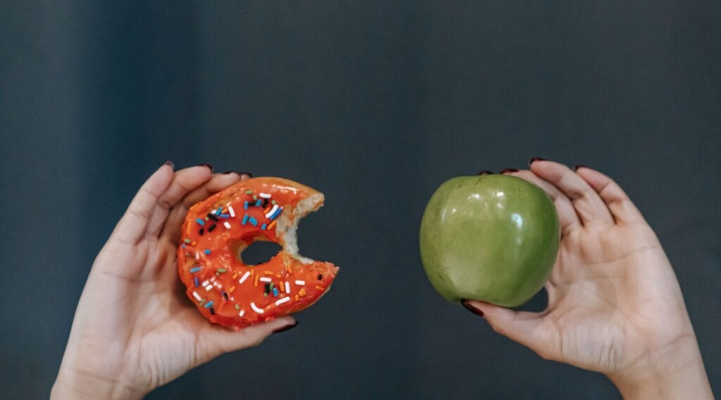 A hand holding a doughnut beside a hand holding a green apple, representing ultra-processed food compared with whole food.